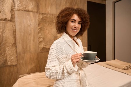 Téléchargez les photos : Jeune femme en peignoir sourit, tenant une tasse et attendant dans la salle de spa - en image libre de droit