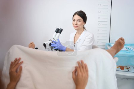 Téléchargez les photos : Gynécologue souriante femelle est assis et faire un examen de l'utérus dame à l'aide d'un microscope - en image libre de droit