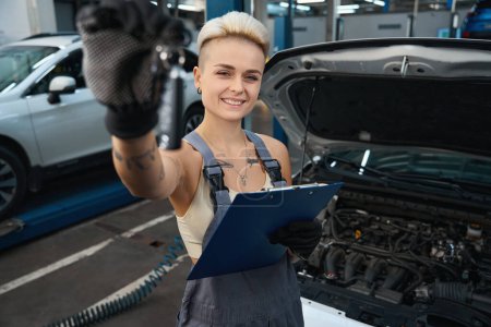 Photo for Young woman mechanic in gloves holding clipboard and looking at camera with happy facial expression at maintenance station - Royalty Free Image