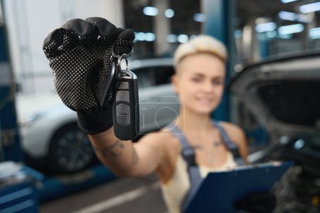 Téléchargez les photos : Vue rapprochée de la main du mécanicien féminin en gant montrant la clé du véhicule à la caméra sur fond flou du service de voiture - en image libre de droit