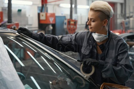 Foto de Mecánica femenina concentrada en el trabajo cinta adhesiva adhesiva uniforme para parabrisas en el servicio de reparación de automóviles - Imagen libre de derechos