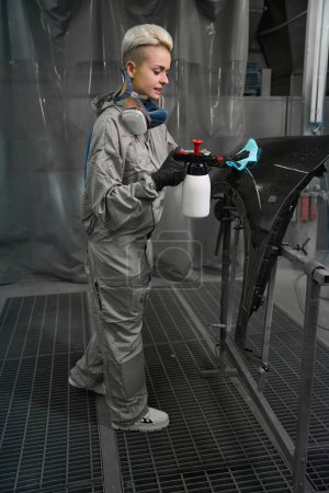 Téléchargez les photos : Jolie femme technicienne de désinfection dans les vêtements de travail et respirateur autour du lavage du cou élément automobile dans l'atelier de réparation automobile - en image libre de droit