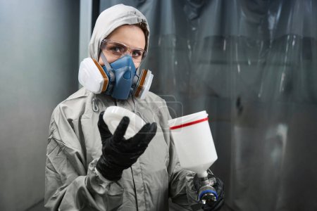 Foto de Mujer trabajadora caucásica en ropa protectora mirando la cámara mientras verifica la cantidad de pintura en el aerógrafo en el garaje - Imagen libre de derechos
