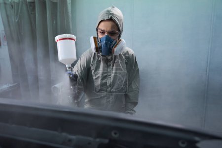 Foto de Trabajadora responsable en ropa protectora y respirador rociando pintura en garaje de servicio - Imagen libre de derechos