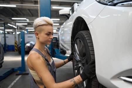 Téléchargez les photos : Femme maître avec une coupe de cheveux élégant examine une roue de voiture, un bon éclairage à l'intérieur - en image libre de droit