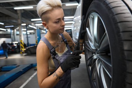 Téléchargez les photos : Femme mécanicien automobile en salopette de travail vérifie une roue de voiture, elle utilise un outil spécial - en image libre de droit