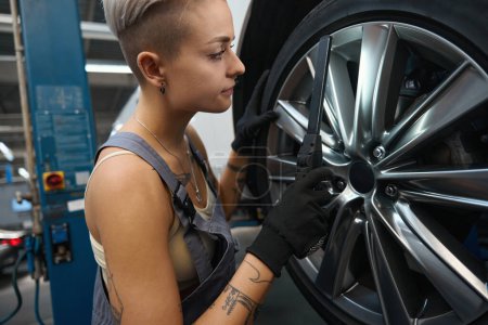 Téléchargez les photos : Jeune femme mécanicien automobile travaille avec une roue de voiture, elle utilise un outil spécial - en image libre de droit