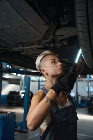 Photo for Woman foreman carries out repair work under a car raised on a lift, a woman uses a special lamp - Royalty Free Image