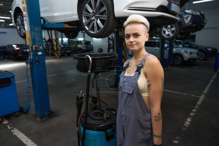 Foto de Mujer reparador de automóviles se para con un dispositivo para el drenaje de aceite, junto a un coche en un ascensor especial - Imagen libre de derechos