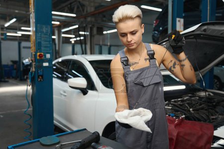 Téléchargez les photos : Femme en salopette de travail vérifie le niveau d'huile dans la voiture, l'artisan utilise un outil spécial - en image libre de droit