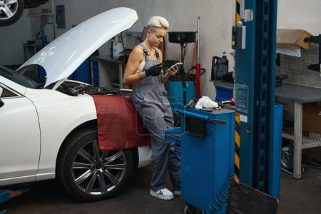 Téléchargez les photos : Jeune femme dans un atelier de voiture se tient près d'une voiture avec le capot levé, elle a un téléphone dans ses mains - en image libre de droit