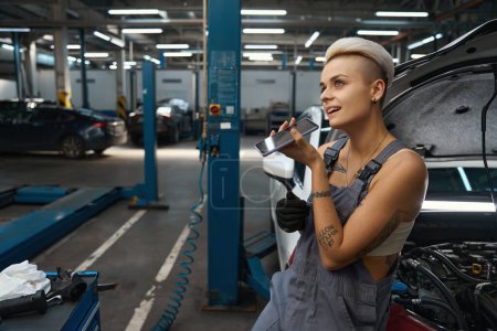 Téléchargez les photos : Jeune femme avec une coupe de cheveux courte est debout parmi les voitures dans l'atelier de réparation automobile, une artisan parle au téléphone - en image libre de droit