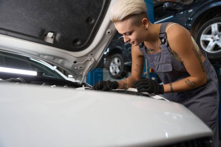 Photo for Young female in work overalls repairs a car engine, a craftswoman uses protective gloves - Royalty Free Image
