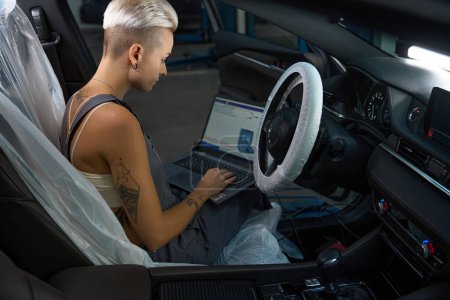 Photo for Car mechanic in work overalls conducts computer diagnostics of a car, a woman uses a laptop - Royalty Free Image