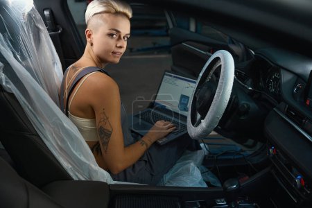 Photo for Woman auto mechanic sits in car interior protected by covers with laptop, female with a tattoo and in work overalls - Royalty Free Image