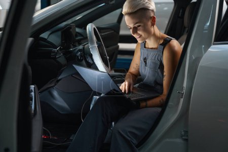 Photo for Repairwoman with a stylish short haircut sits in the car with a laptop, a woman in overalls - Royalty Free Image