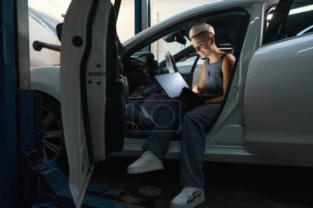 Photo for Young repairwoman sitting in a car interior with a laptop and typing, the female has a stylish short haircut - Royalty Free Image