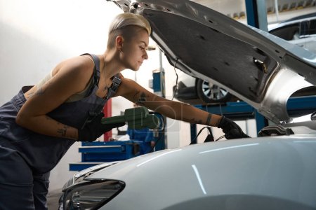 Téléchargez les photos : Mécanicien automobile travaille sous le capot d'une voiture avec un scanner spécial, une femme en salopette de travail - en image libre de droit