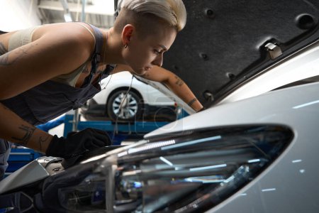 Photo for Young woman auto mechanic checks the engine of a car, female with a tattoo and in work overalls - Royalty Free Image