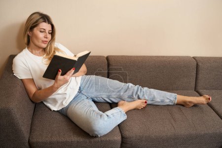 Téléchargez les photos : Belle jeune femme en vêtements décontractés livre de lecture tout en étant couché sur le canapé dans la chambre moderne - en image libre de droit