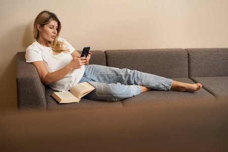 Téléchargez les photos : Belle femelle en vêtements décontractés tenant le téléphone portable dans les mains et se détendre sur le canapé dans le salon des appartements - en image libre de droit