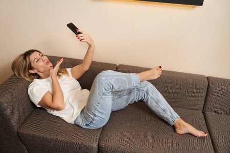 Téléchargez les photos : Jolie femelle allongée sur le canapé et croisée ses jambes tout en soufflant baiser lors d'un appel vidéo par mobile à l'intérieur - en image libre de droit