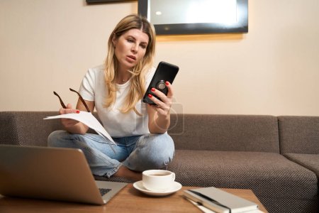Téléchargez les photos : Femme attrayante en vêtements décontractés assis sur le canapé et en utilisant mobile pendant les missions sur ordinateur portable à l'intérieur - en image libre de droit