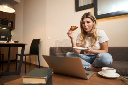 Téléchargez les photos : Assis jeune femme aux cheveux longs manger croissant avec du café tout en regardant ordinateur portable dans la cuisine - en image libre de droit