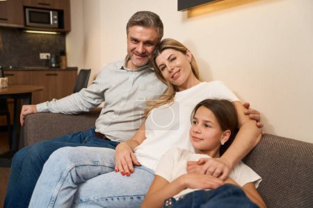 Foto de Esposo y esposa con hija sentados en el sofá y posando en la cámara en la habitación moderna - Imagen libre de derechos