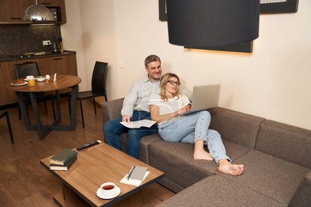 Photo for Handsome man sitting on cozy sofa and looking at laptop of wife which is on her lap in apartment - Royalty Free Image