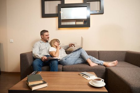 Foto de Vista lateral del hombre relajado sentado junto a la esposa pensativa en gafas con cuaderno en sus rodillas en la habitación - Imagen libre de derechos