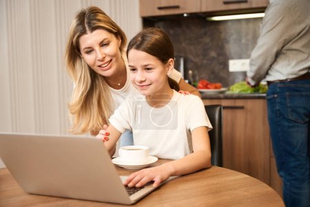 Téléchargez les photos : Mère souriante et jolie fille sont assis à la table avec un ordinateur portable dans la cuisine dans la chambre d'hôtel et thé en nature. L'homme se tient en arrière-plan et cuisine - en image libre de droit