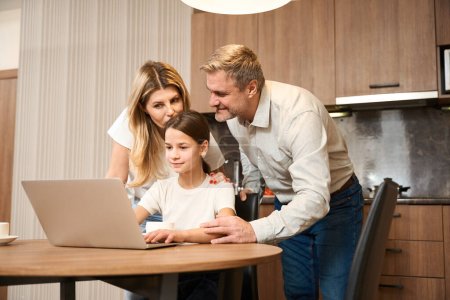 Photo for Little girl is sitting at table with laptop in the kitchen and drinking tea. Smiling woman is standing behind, kissing and huging her kid. Happy man is standing next to and holding the girl hand - Royalty Free Image