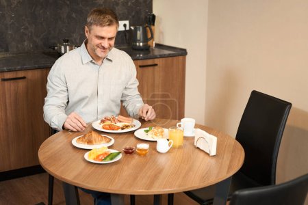 Téléchargez les photos : Homme adulte est assis à table dans la cuisine et prendre le petit déjeuner beaucoup de délicieux repas à l'hôtel - en image libre de droit
