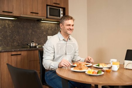 Téléchargez les photos : Un gars souriant est assis à table dans la cuisine et prend le petit déjeuner beaucoup de délicieux repas à l'hôtel - en image libre de droit