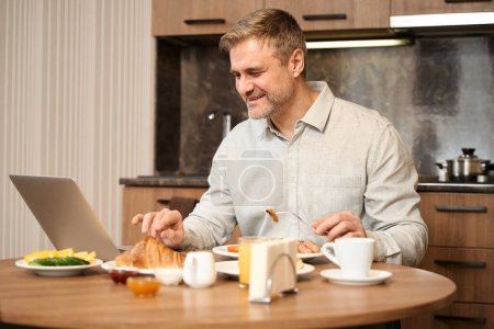Foto de Hombre sonriente está sentado en la mesa, en línea trabajando con el ordenador portátil y comer deliciosa comida en la cocina en la habitación de hotel - Imagen libre de derechos