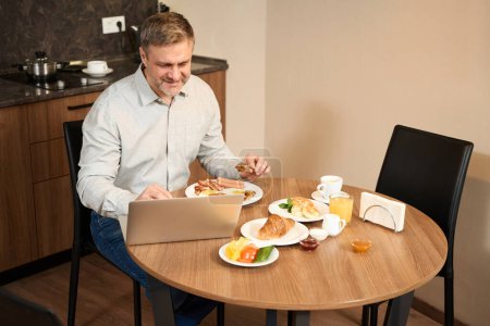 Téléchargez les photos : Souriant mâle est assis à la table et en ligne de travail avec ordinateur portable dans la cuisine et de manger de délicieux aliments - en image libre de droit
