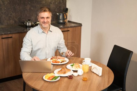 Téléchargez les photos : Souriant mâle est assis à la table avec ordinateur portable dans la cuisine et de manger de délicieux aliments - en image libre de droit