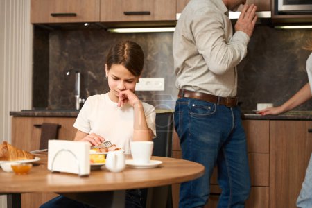 Photo for Upset kid female is sitting at the table in the kitchen, eating and drinking. Her parents are in the background discussing something - Royalty Free Image