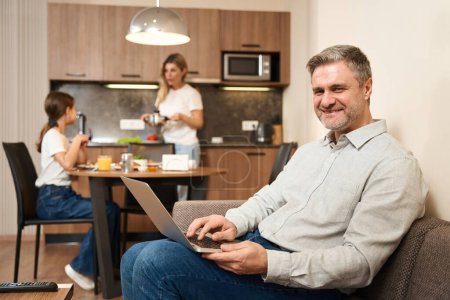 Téléchargez les photos : Un homme souriant est assis sur le canapé avec un ordinateur portable. Femme et petite fille préparent le petit déjeuner en arrière-plan - en image libre de droit