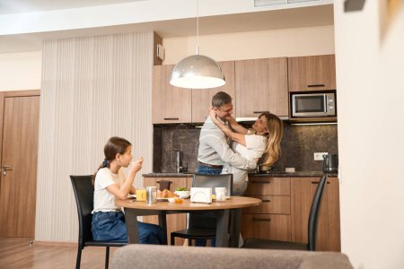 Foto de La niña está sentada en la mesa, comiendo y bebiendo té. Sus padres y bailando en la cocina en la habitación del hotel - Imagen libre de derechos