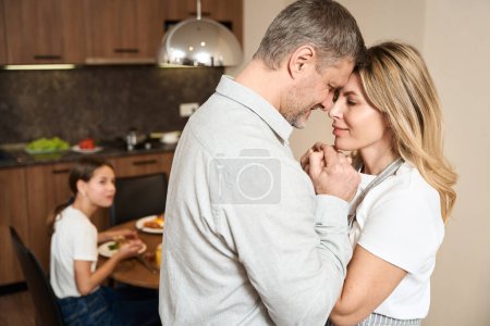 Téléchargez les photos : Agréable gars et belle dame se tiennent la main et se regardent avec amour dans la cuisine. Petite fille est assise à table et prend le petit déjeuner en arrière-plan - en image libre de droit