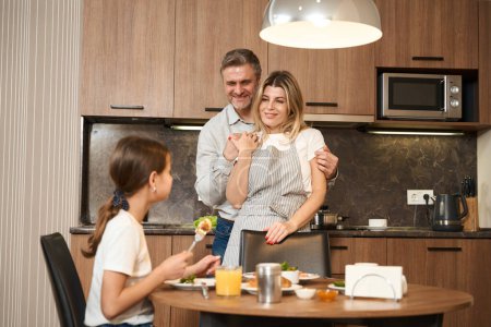Photo for Pleasant man and smiling woman are standing, hugging and looking at daughter. Little girl is sitting at table and having breakfast in hotel room - Royalty Free Image