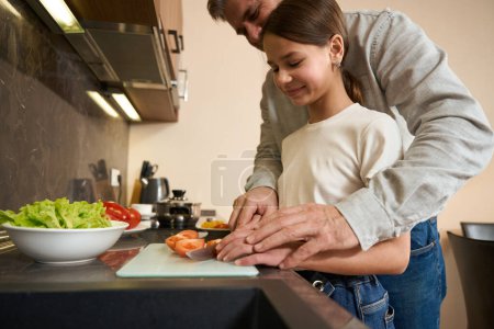Téléchargez les photos : Père aide la fille à couper des tomates pour le petit déjeuner dans la cuisine. Plaques avec des produits sont sur la surface de la cuisine - en image libre de droit