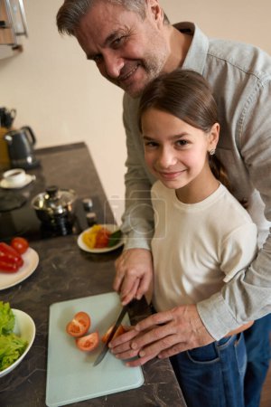 Photo for Happy father is helping the girl to cut tomatoes for breakfast in the kitchen. Plates with products are on the kitchen surface - Royalty Free Image
