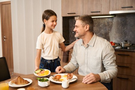 Téléchargez les photos : Un homme souriant est assis à la table avec de la nourriture dans la cuisine et regardant l'enfant. Jolie fille met la plaque de nourriture sur la table et souriant en arrière - en image libre de droit