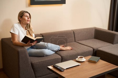 Foto de Agradable dama está sentada sola en el sofá, relajarse y leer un libro en la habitación del hotel - Imagen libre de derechos