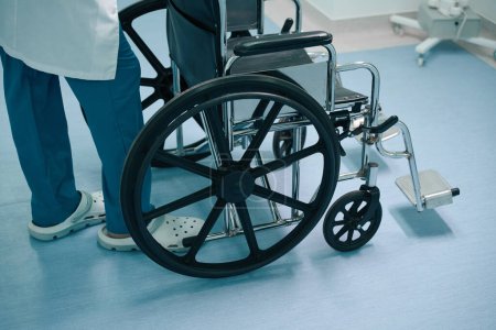Photo for Uniformed nurse stands in a hospital corridor with a wheelchair, the chair has large and small wheels - Royalty Free Image