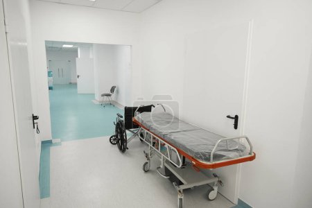 Téléchargez les photos : Lit postopératoire mobile et un fauteuil roulant dans le couloir de l'hôpital, la chambre est lumineuse et propre - en image libre de droit