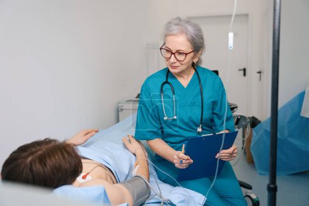Foto de Paciente joven está en tratamiento en un centro de cardiología, una doctora escribe recomendaciones - Imagen libre de derechos